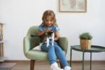 A young girl sits on an armchair, gently holding a kitten beside a potted plant in a cozy room.