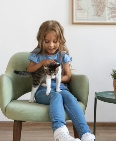 A young girl sits on an armchair, gently holding a kitten beside a potted plant in a cozy room.